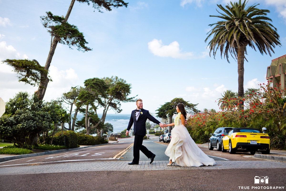 A couple in formal attire walks hand in hand across a street with palm trees and cars, overlooking the ocean.