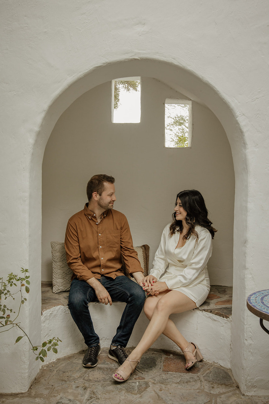 A man and woman sit together in an alcove, holding hands and smiling at each other, with three small windows behind them.