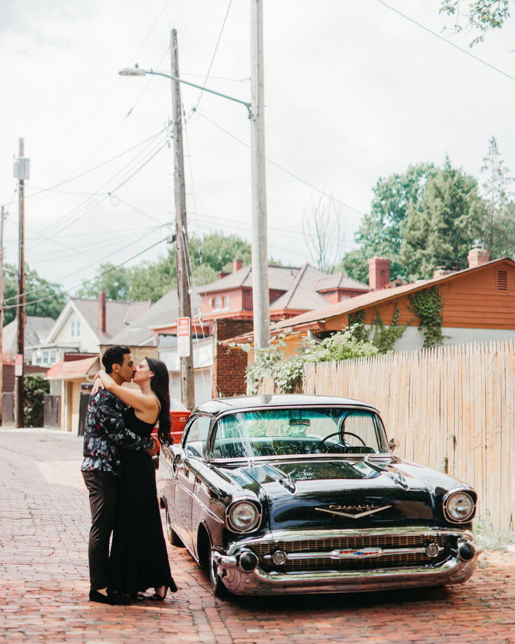 A couple embraces beside a vintage black Chevrolet on a cobblestone street, surrounded by houses and utility poles.