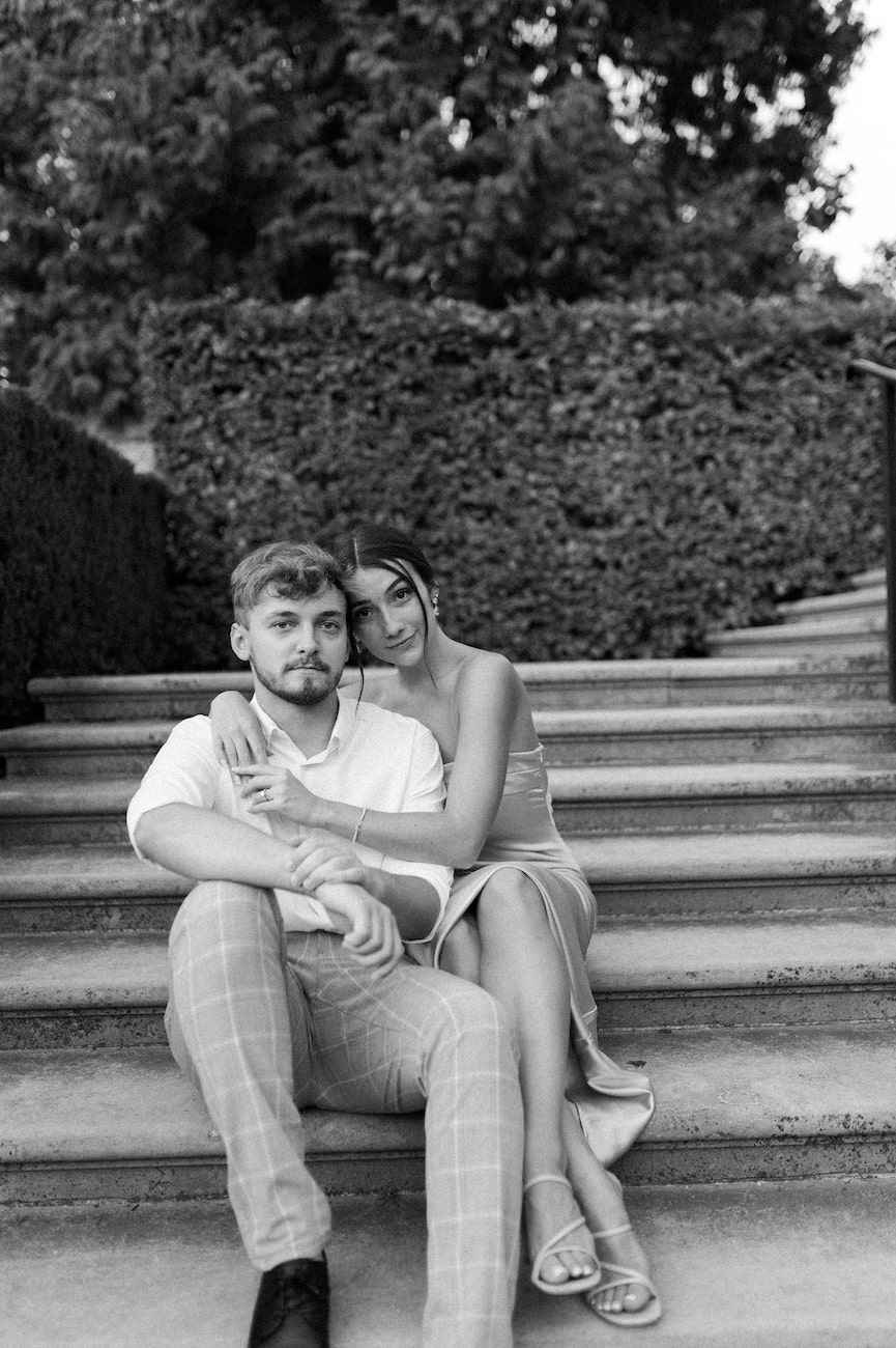 A couple sits on outdoor steps; the woman leans against the man with her arms around him, and both look towards the camera. Black and white image.