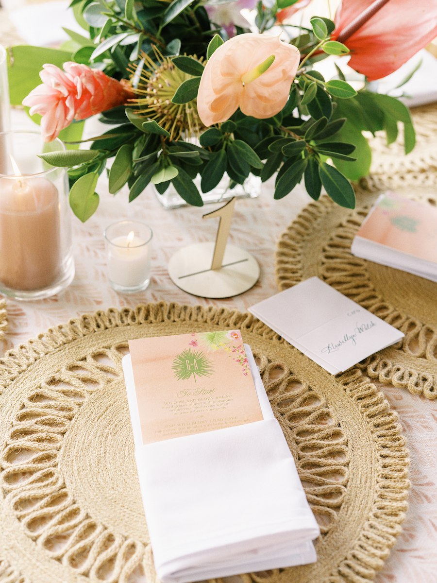 Floral centerpiece with pink anthuriums on a table, set with woven placemats, a menu, napkin, candles, and a small card marked "Table 1".