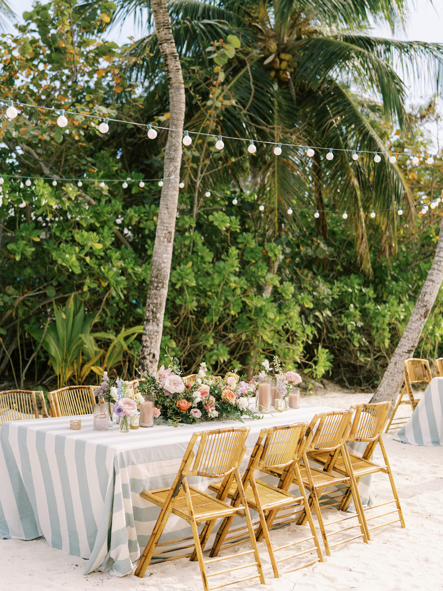 A long table with a striped tablecloth is set on sand, surrounded by bamboo chairs. It is decorated with flower arrangements. String lights hang above between palm trees in the background.