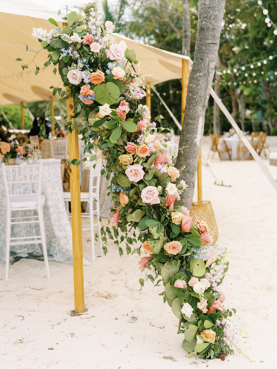 Floral arrangement of pink, orange, and white roses with green leaves decorates a tree trunk at an outdoor event with chairs and tables under a canopy.