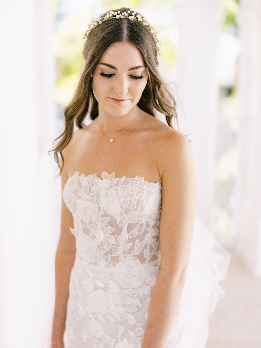 A woman in a white floral lace strapless gown and floral headpiece stands indoors with her eyes downcast.