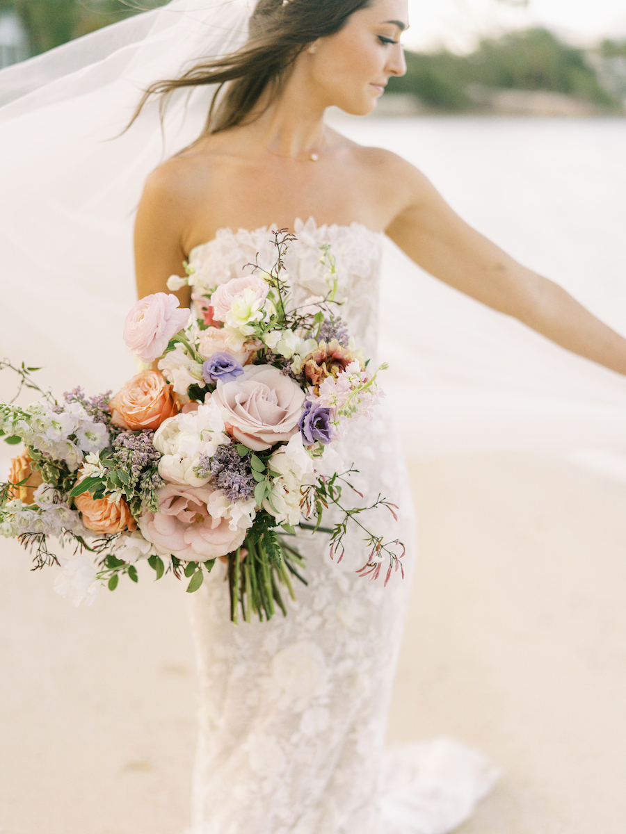 Bride in a strapless lace gown holding a large bouquet of pastel flowers on a beach with wind blowing her veil.