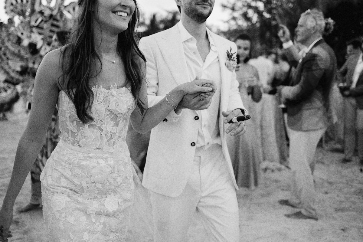 A couple wearing wedding attire walks together during a celebration, surrounded by guests in an outdoor setting.