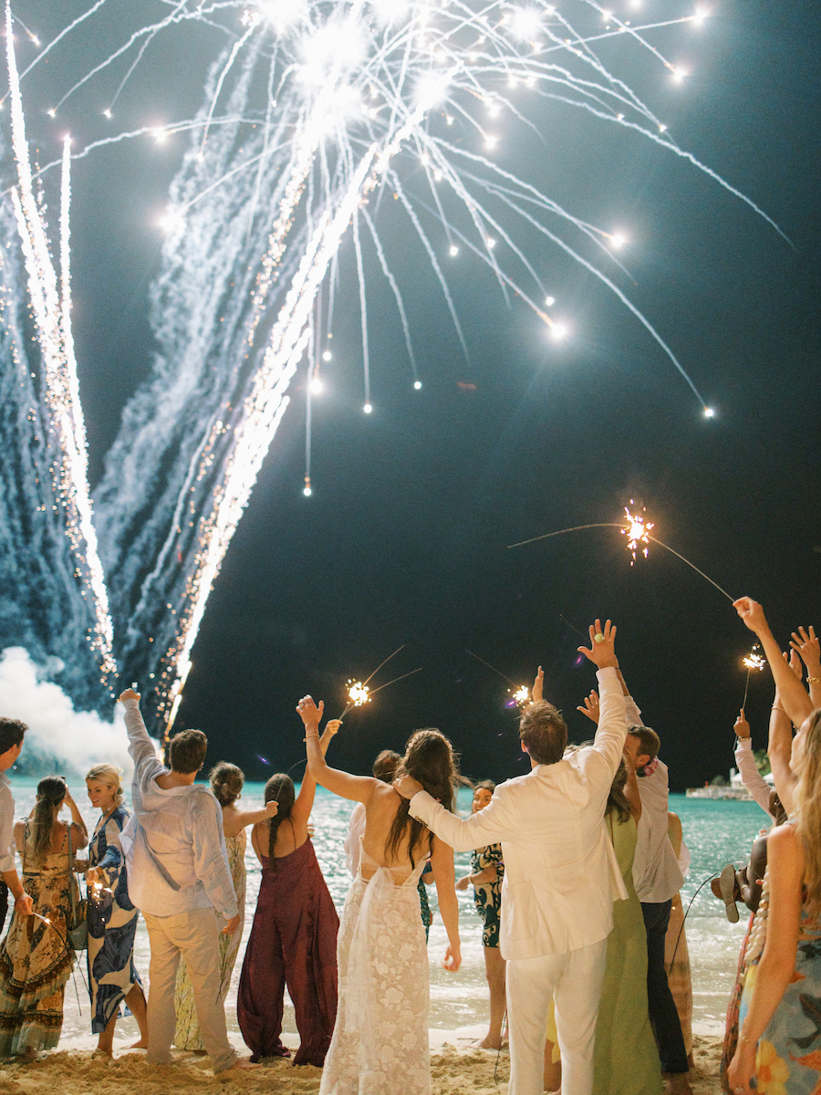 A group of people celebrate on a beach at night, holding sparklers and watching fireworks in the sky.