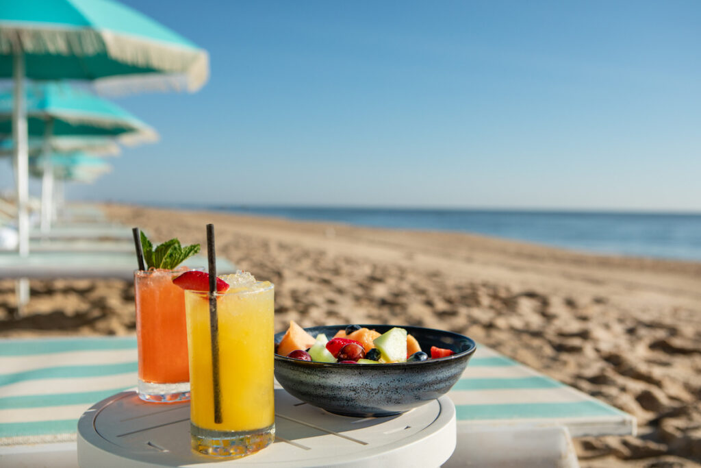 Two colorful cocktails and a bowl of fruit on a beachside table with sand and ocean in the background.