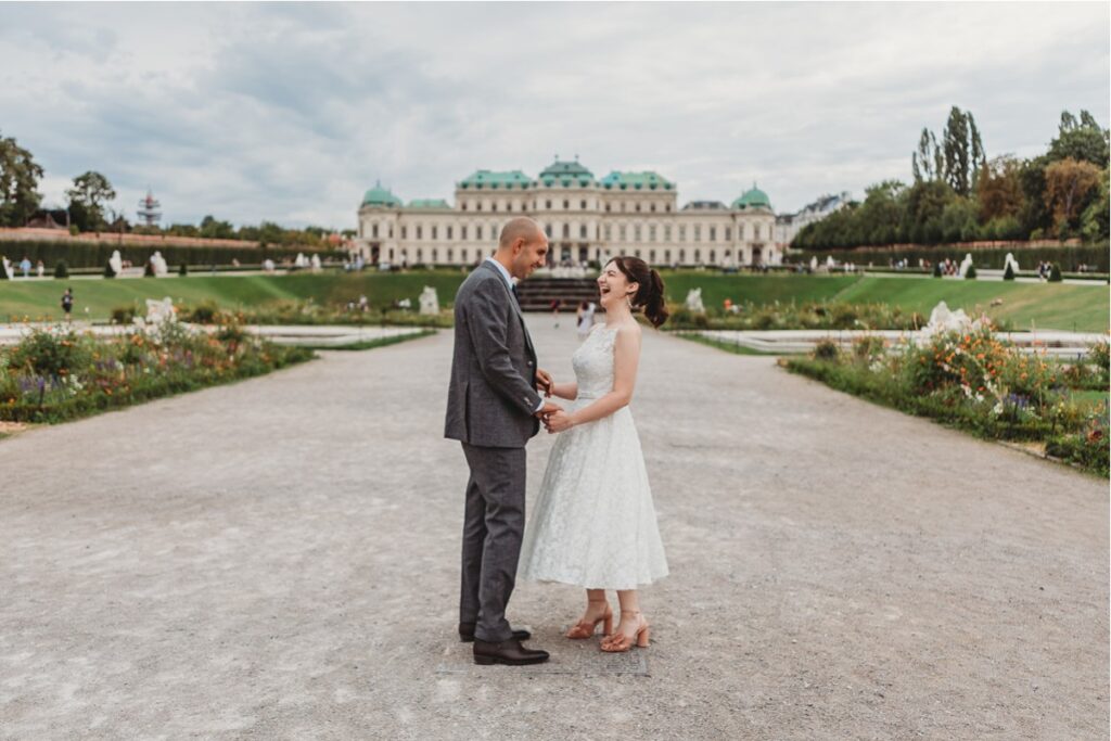 A couple stands holding hands on a gravel path, with a large building and gardens in the background.