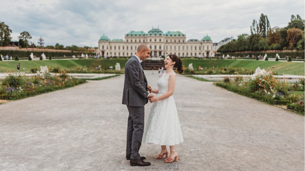 A couple stands holding hands on a gravel path, with a large building and gardens in the background.