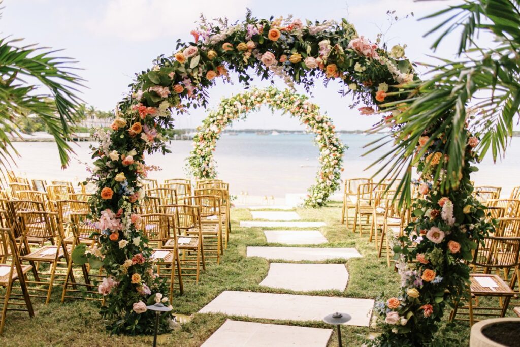 Tropical outdoor wedding setup with floral arches over a grassy aisle, rows of wooden chairs, and a beach backdrop.