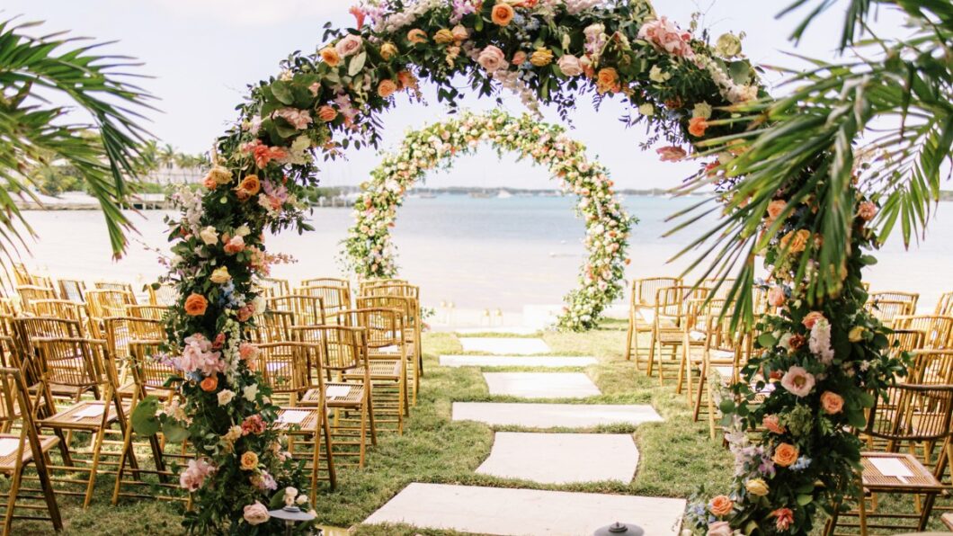 Tropical outdoor wedding setup with floral arches over a grassy aisle, rows of wooden chairs, and a beach backdrop.