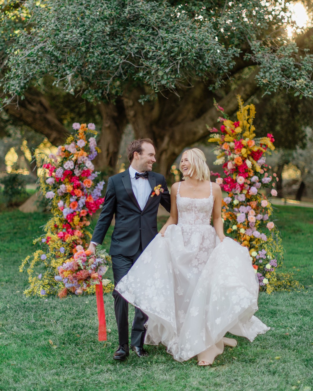 A couple in formal attire smiling and holding hands, standing in front of vibrant floral arrangements in a lush garden setting.