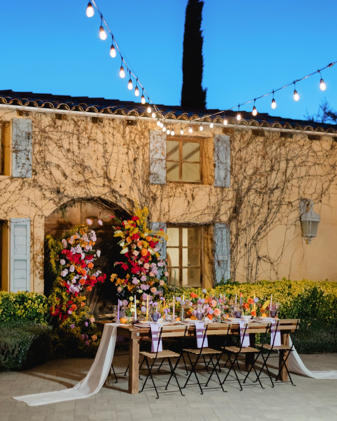 Outdoor dining setup with decorated table, lit candles, and floral arrangement under string lights. A rustic building with ivy and blue shutters is in the background.