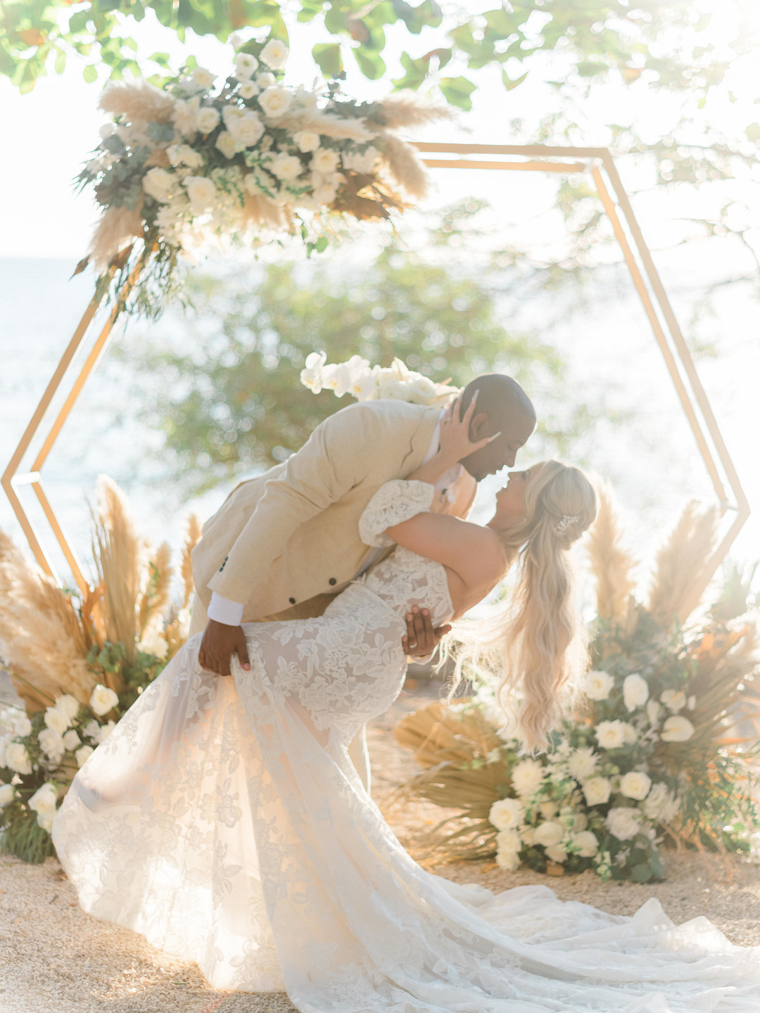 A couple shares a kiss during their wedding ceremony in front of a hexagonal floral arch with ocean in the background.