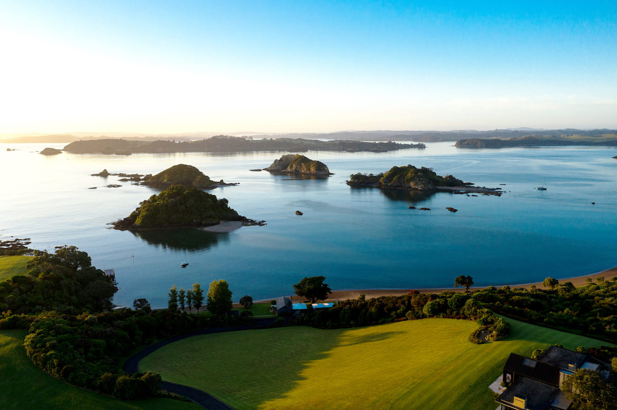 Aerial view of a coastal New Zealand with small islands surrounded by calm blue water, a green field in the foreground, and a clear sky above.