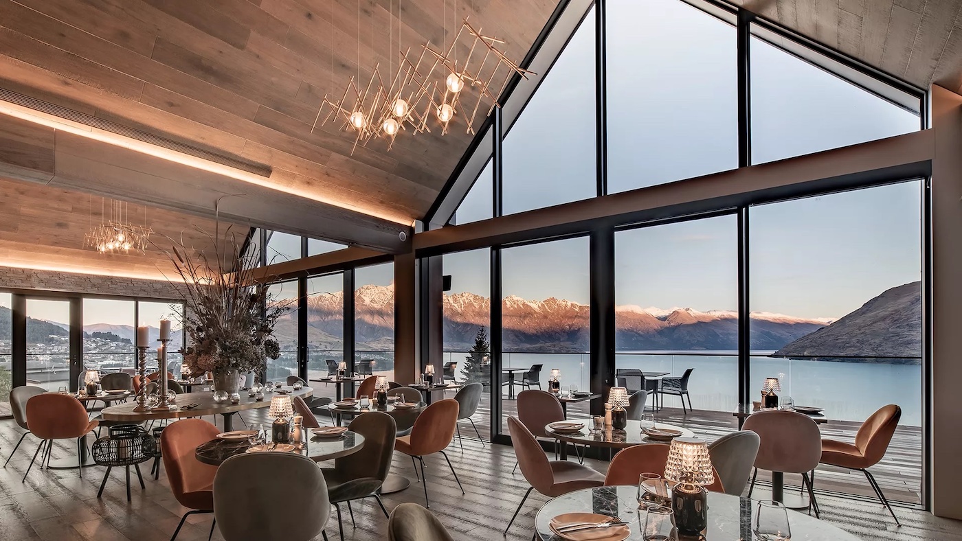 Modern dining area with large windows, offering a panoramic view of a lake and mountains at sunset. Tables are set for service with elegant decor and lighting.