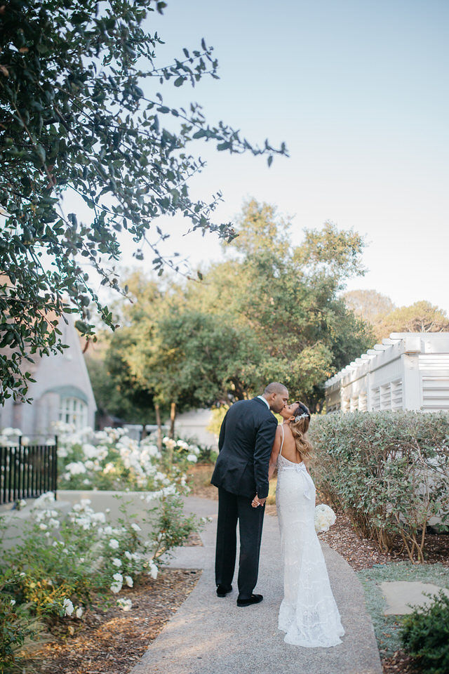 A bride and groom kiss while walking along a garden path, surrounded by greenery and white flowers.