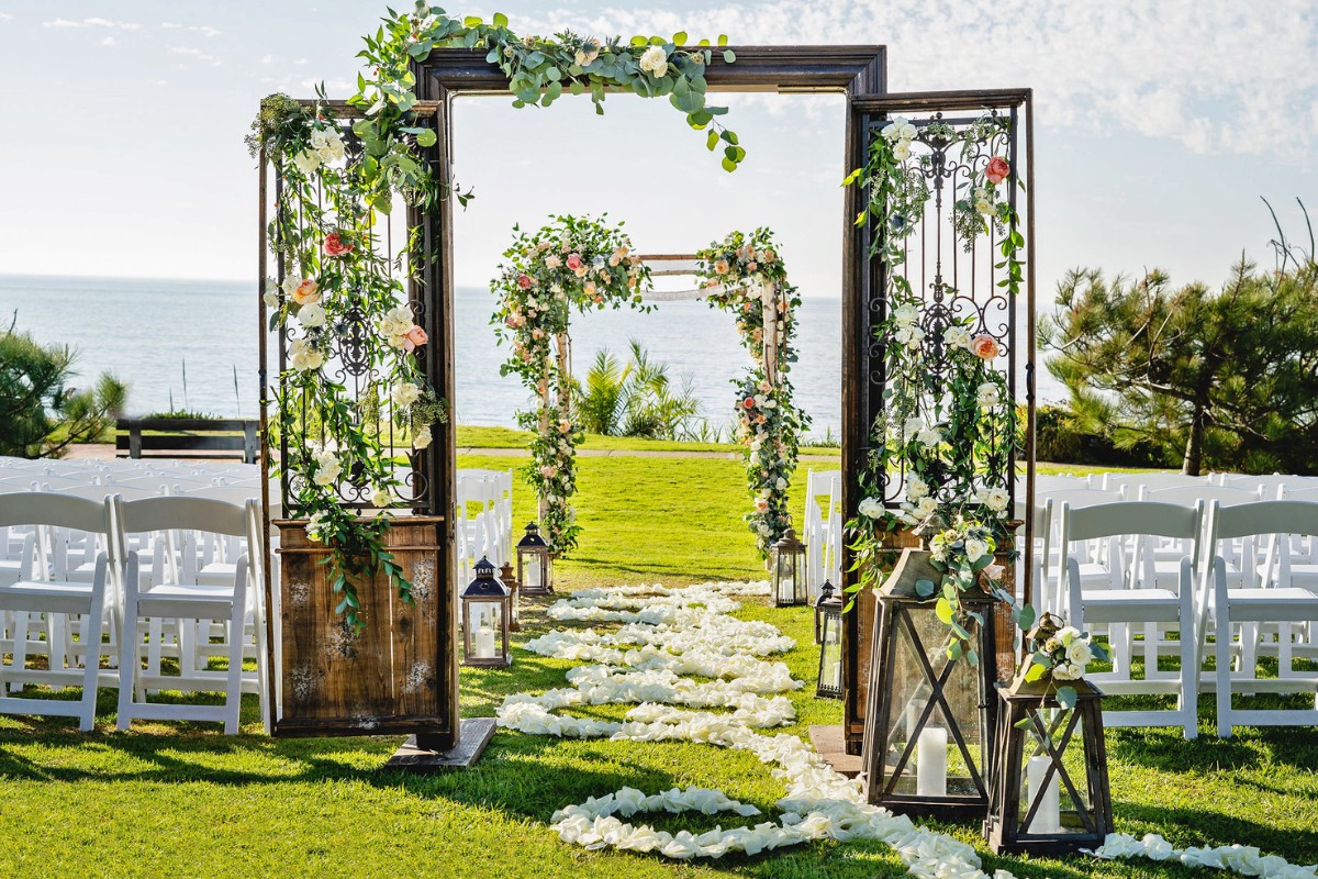 Outdoor wedding setup with white chairs facing an ocean view. An archway adorned with flowers and greenery leads to an altar. Rose petals line the aisle, and lanterns are placed along the path.