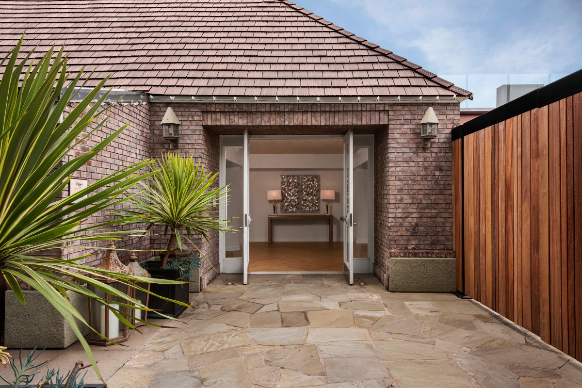 Brick building entrance with double glass doors open, surrounded by stone tiles and decorative plants. Wooden fence on the right.