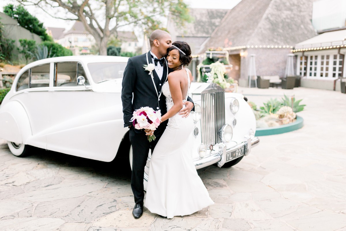 A groom in a black tuxedo kisses a bride in a white dress, holding pink and white flowers, beside a vintage white car on a stone pavement.