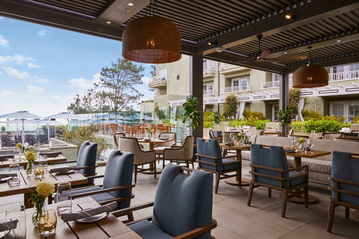 Outdoor restaurant patio with wooden tables and chairs, set with glasses and napkins. View of the ocean and nearby buildings in the background.