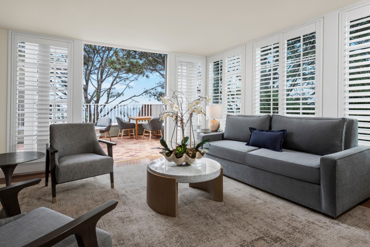 A modern living room with gray furniture, a round table with an orchid centerpiece, and large windows leading to a patio with outdoor seating and a view of trees.