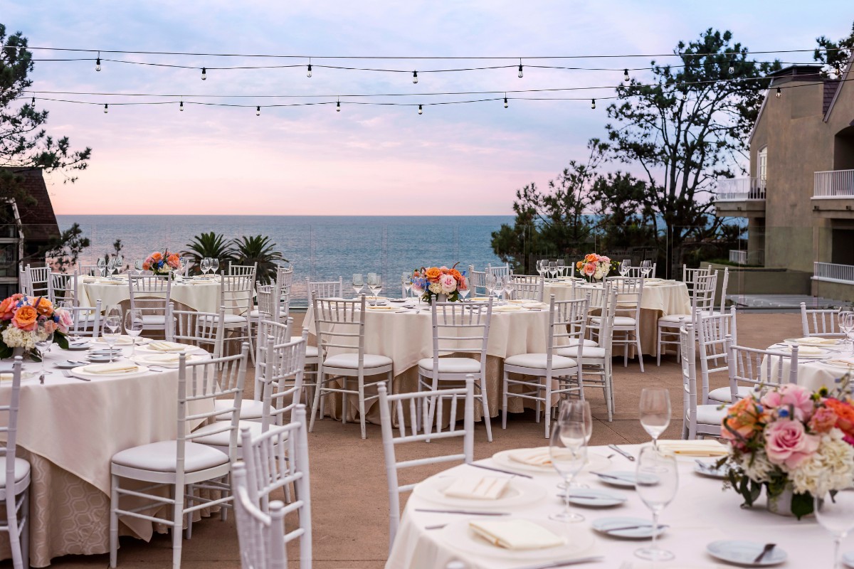 Outdoor event setup with round tables and white chairs on a patio overlooking the ocean. Each table has floral centerpieces. String lights are hanging overhead.