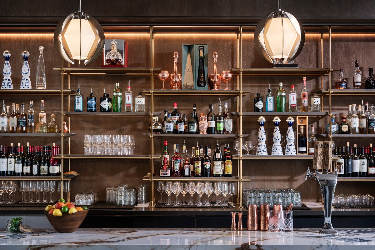 Bar interior with a variety of liquor bottles neatly arranged on shelves, glasses evenly placed, two geometric pendant lights overhead, and a bowl of fruits on the marble counter.