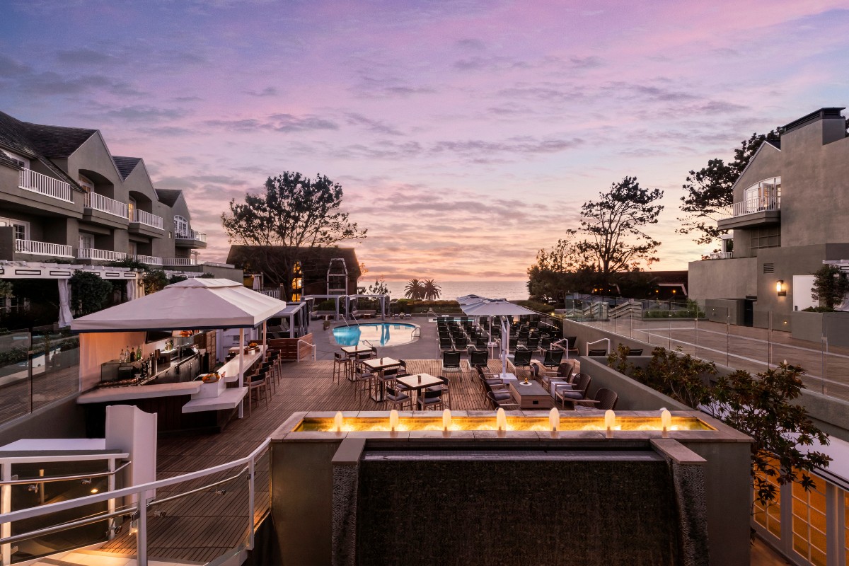 A luxury resort terrace at sunset features a pool, lounge chairs, tables, umbrellas, and trees, with the sky displaying shades of purple and orange.