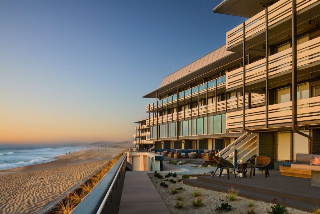 Modern beachfront building with large windows overlooking a sandy beach and ocean at sunset, featuring outdoor seating areas and a boardwalk.