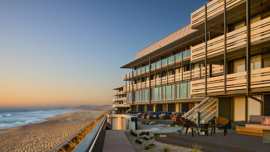 Modern beachfront building with large windows overlooking a sandy beach and ocean at sunset, featuring outdoor seating areas and a boardwalk.