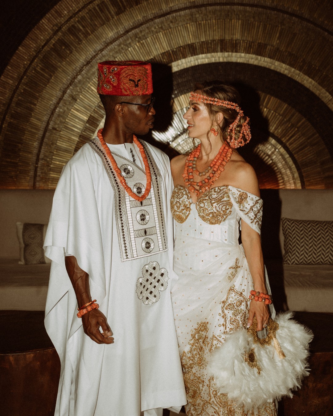 A couple in traditional attire stand facing each other, wearing ornate white and gold garments with red beaded accessories. The background features a decorative, circular metallic wall.