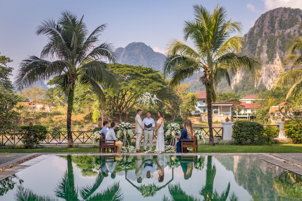 A couple is getting married outdoors near a pool, surrounded by palm trees, mountains, and a small group of people. The ceremony takes place in a tropical setting under a clear sky.