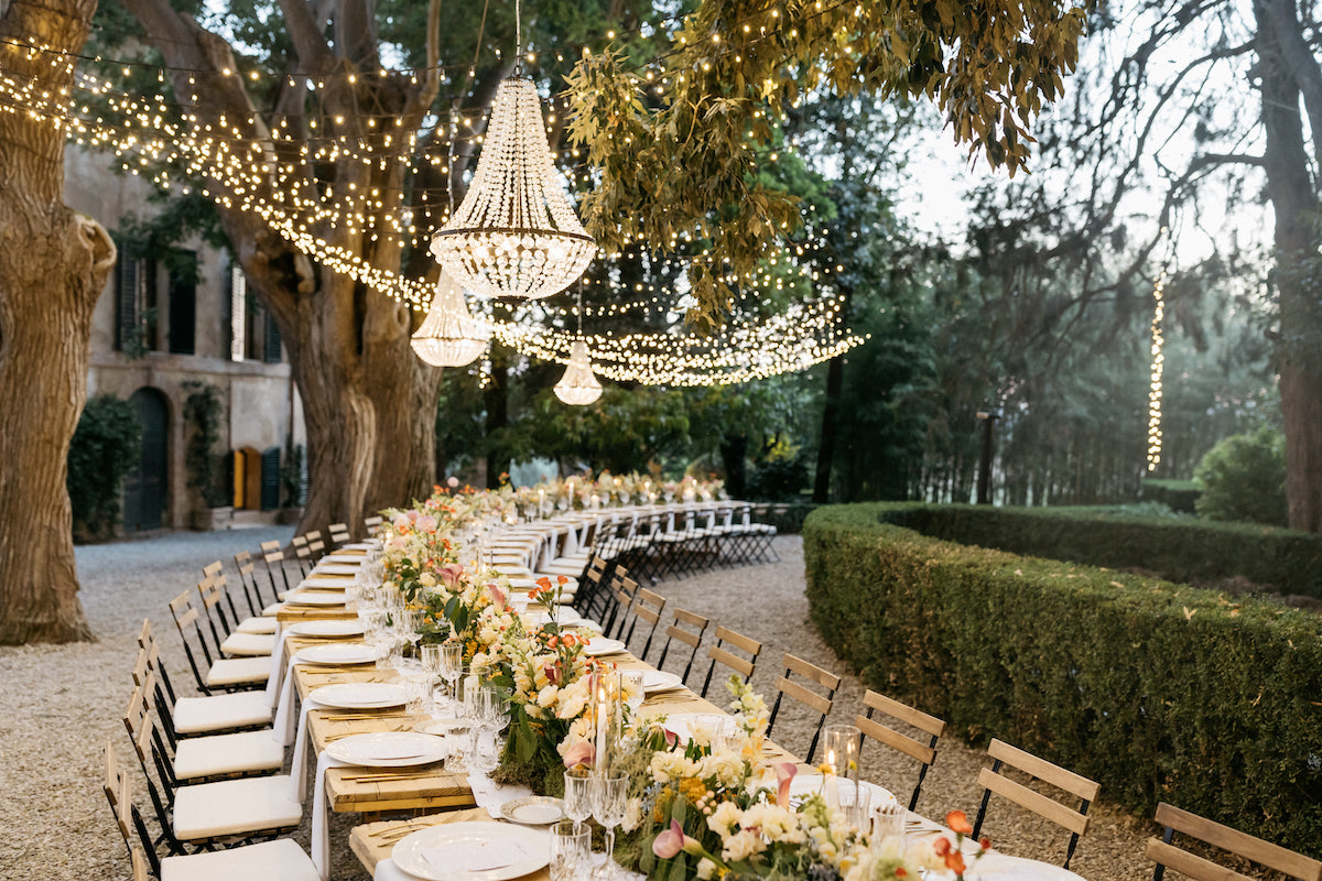 An outdoor long dining table is elegantly set with floral arrangements and tableware under hanging chandeliers and string lights.