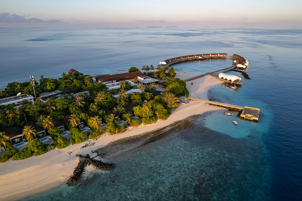 Aerial view of a tropical island resort with lush greenery, sandy beaches, a curved walkway, and structures extending over clear blue water.