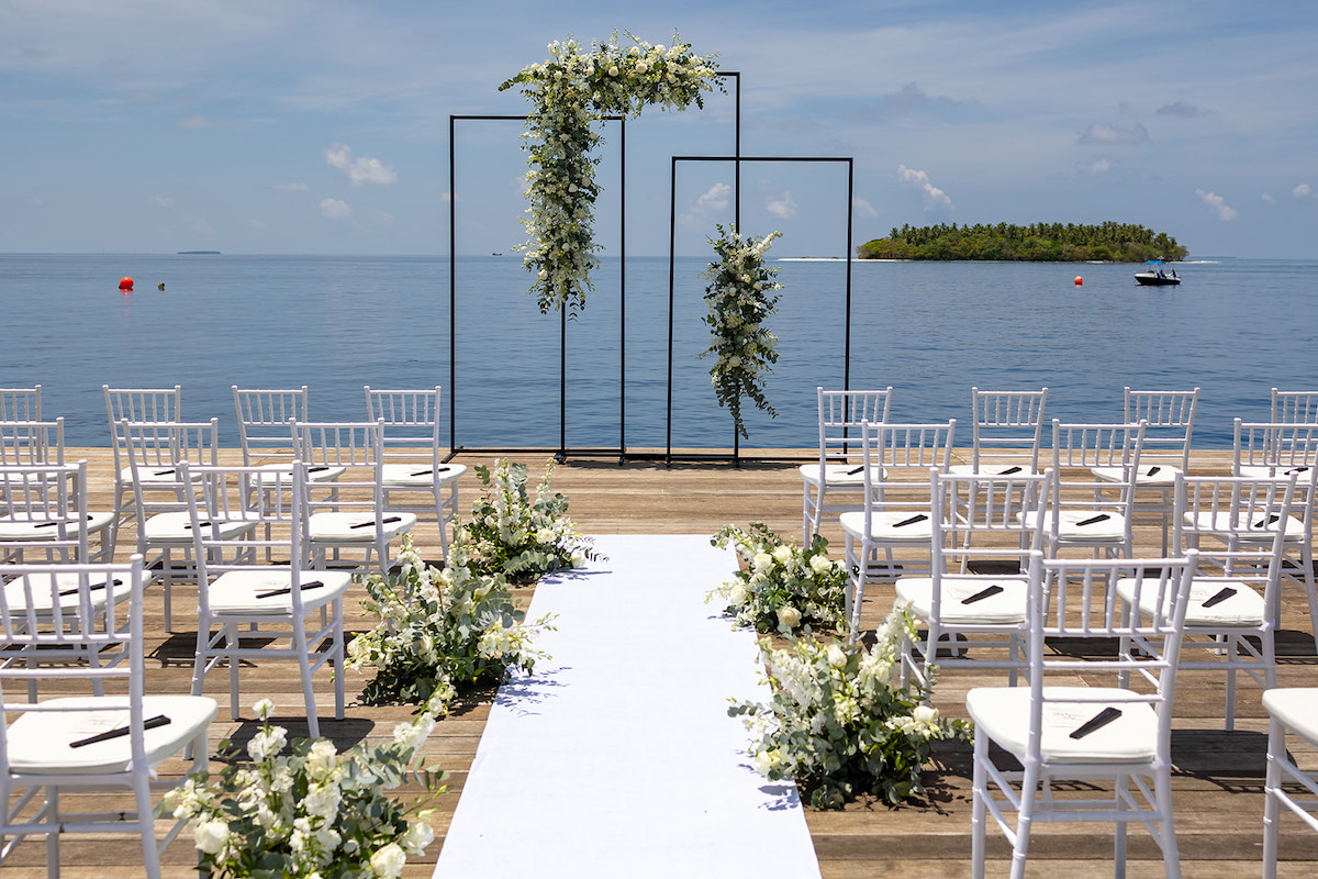 Outdoor wedding setup with white chairs and a floral-decorated aisle facing the ocean. A minimalist arch with greenery stands at the front. An island is visible in the background.