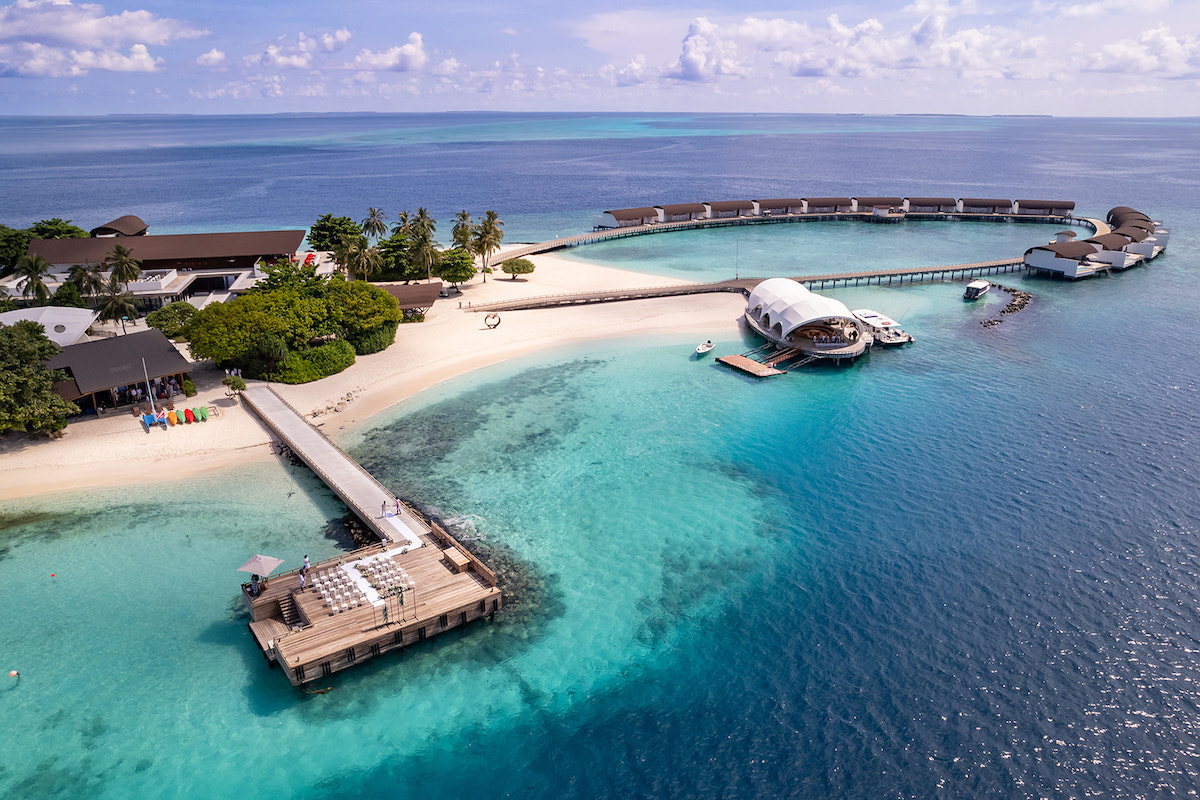 Aerial view of a tropical island resort with overwater villas, a sandy beach, lush greenery, and a long pier extending into clear turquoise waters.