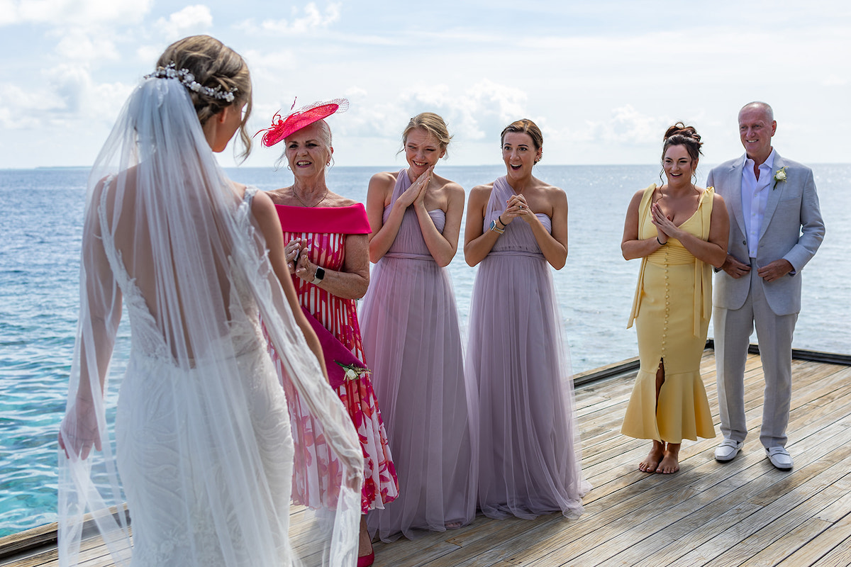 A bride approaches four people on a wooden deck by the sea, with three women in dresses and one man in a light suit.