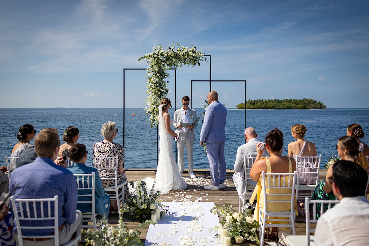 Outdoor wedding ceremony by the sea with a couple standing under an arch adorned with white flowers, officiant in between, guests seated on either side, and an island visible in the background.