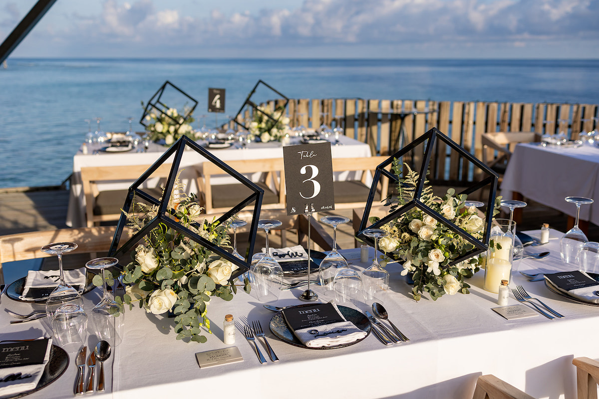 Elegant seaside table setup with geometric centerpieces, white roses, and place settings for an event. Sky and ocean in the background.