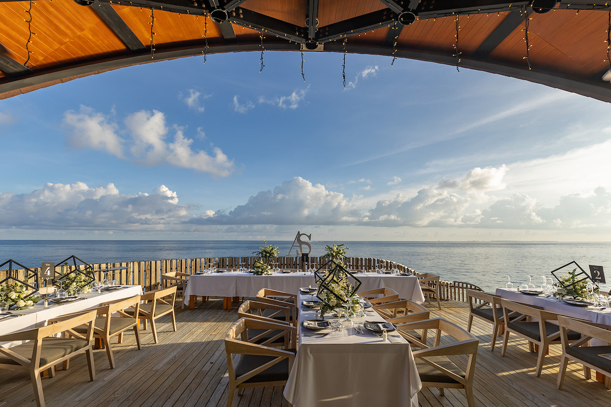 Outdoor dining setup on a wooden deck with tables set for a meal, overlooking a calm ocean under a partly cloudy sky.