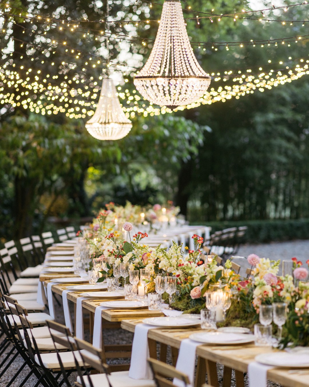 Outdoor dining setup with long wooden tables, floral centerpieces, and white tablecloths. String lights and chandeliers hang above, creating an elegant atmosphere.