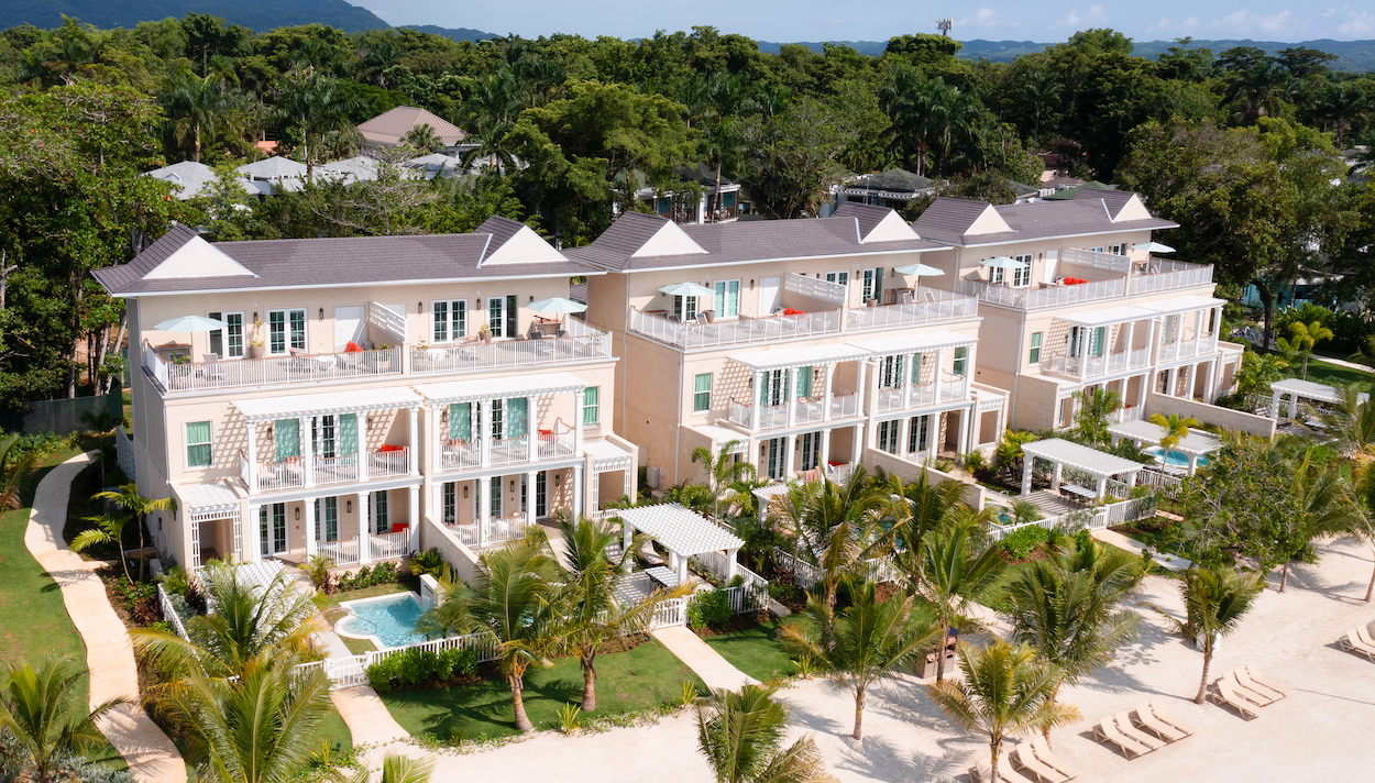 Aerial view of a beachfront resort with three identical beige buildings, multiple balconies, and outdoor seating areas surrounded by palm trees and greenery.