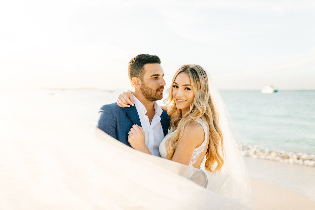 A couple dressed in wedding attire pose on a beach with the ocean in the background. The bride smiles while the groom looks into the distance.