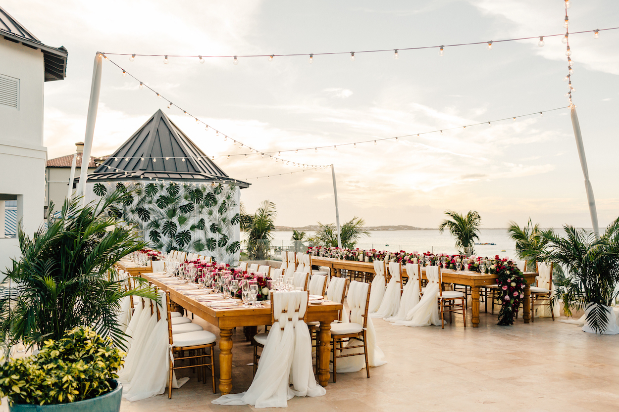 An outdoor wedding reception setup with long wooden tables, white chairs, string lights overhead, and a backdrop of foliage near a waterfront under a cloudy sky at sunset.