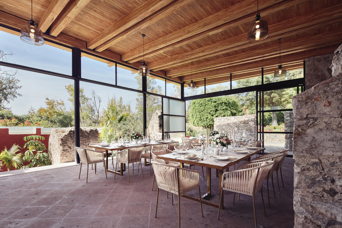 Modern dining area with wooden ceiling, glass walls, and neatly set tables and chairs; natural light entering from large windows overlooking a garden.