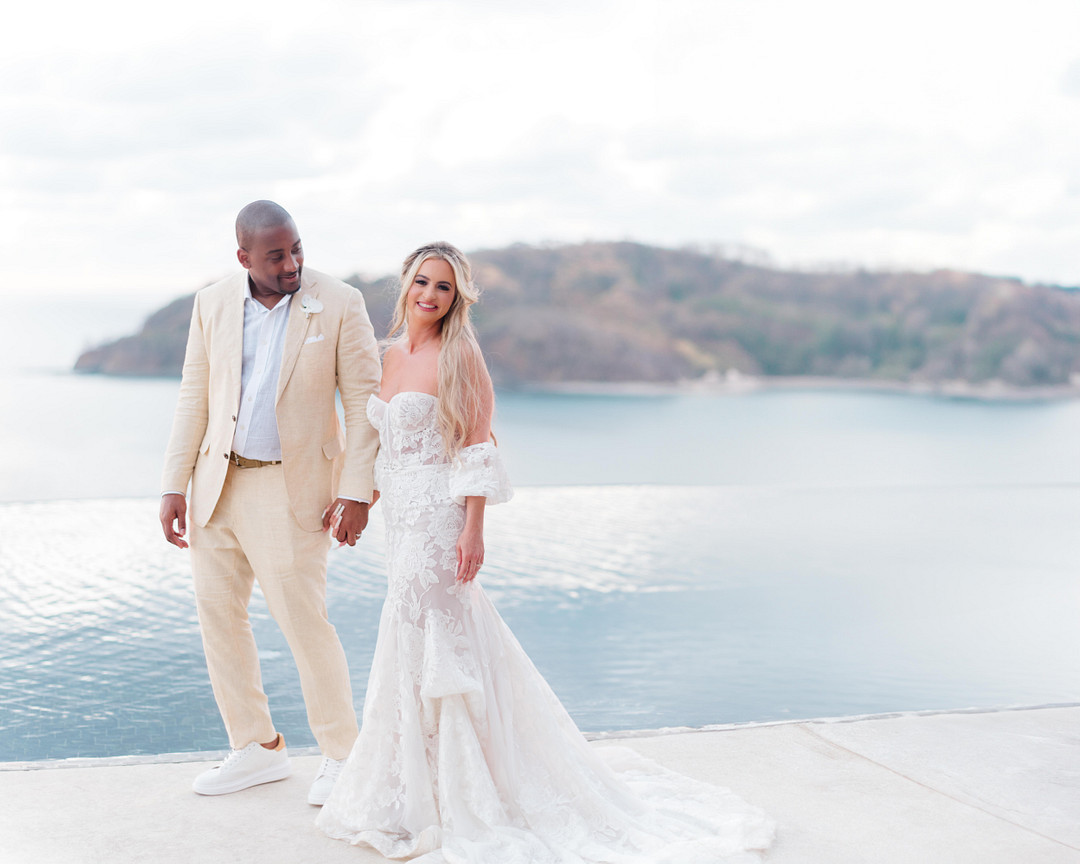 A couple in wedding attire stands by an infinity pool, smiling at each other. The background features a calm seascape and distant island.