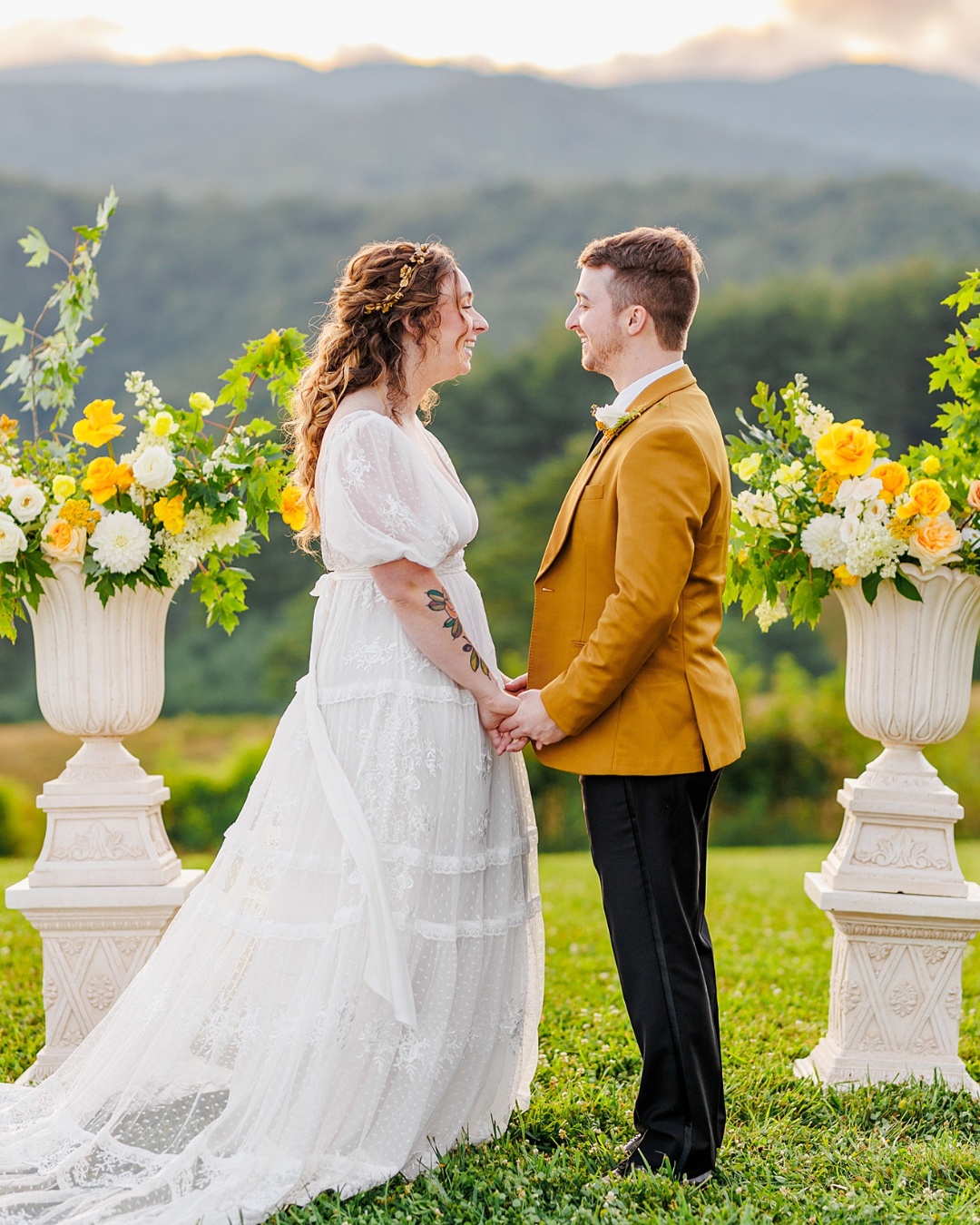 A couple in wedding attire holds hands outdoors, with floral arrangements and green hills in the background.