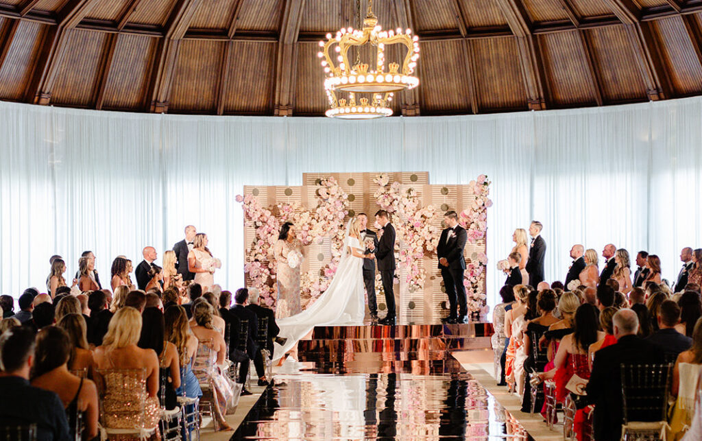 A wedding ceremony inside a domed room with a large chandelier overhead. The couple stands at the altar surrounded by a backdrop of pink flowers, with guests seated on either side.